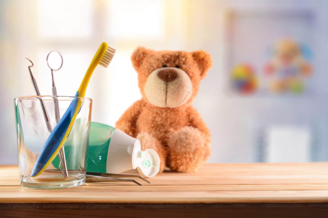 Dentist concept for children with tools in glass cup and teddy on wooden table in room with toys background. Horizontal composition. Front view